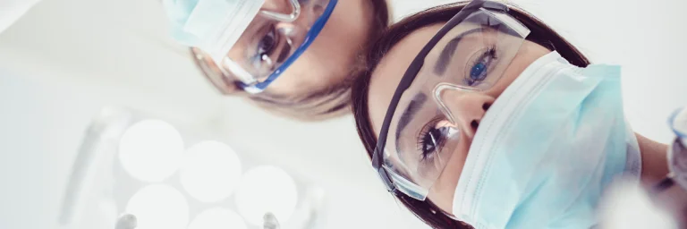 Blickwinkel vom Behandlungsstuhl einer Zahnarztpraxis. Zahnärztin und Zahnmedizinische Fachangestellte schauen auf einen Patienten